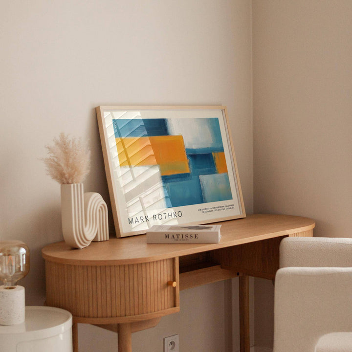 Wide wall art of abstract geometric blocks in blue and orange leaning on a wooden desk beside a white chair