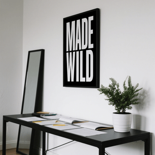 Made Wild art print with white text on black hung above a black desk with a potted fern