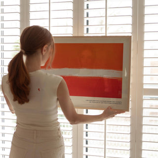 Framed print close-up of horizontal orange and red abstract with white brushstroke held in front of window shutters