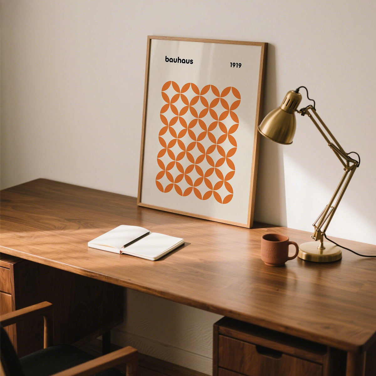 Orange minimalist print leaning on a desk with a brown mug and brass desk lamp