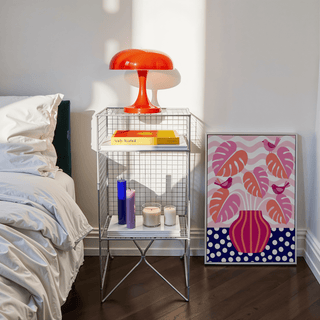 Tropical Plant Wall Art print leaning on the floor with bright pink and orange leaves beside a bedside table with a red lamp