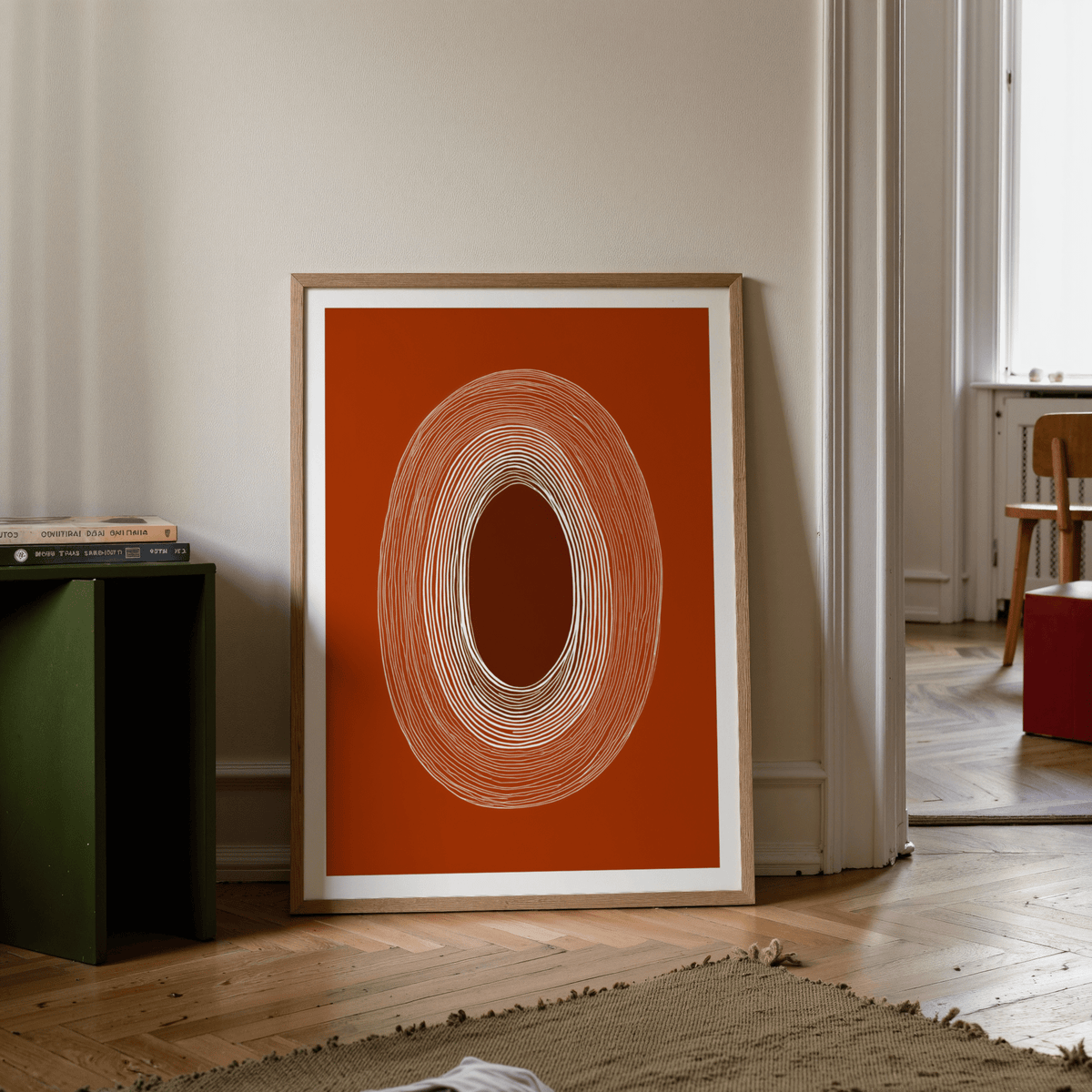 Print leaning on floor with an abstract line drawing poster of concentric oval lines in earthy colours beside a green cabinet