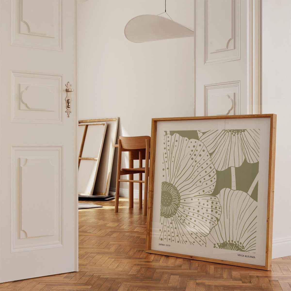 Print leaning on a floor of sage green abstract wall art with stylised poppy flowers near a wooden chair in a room