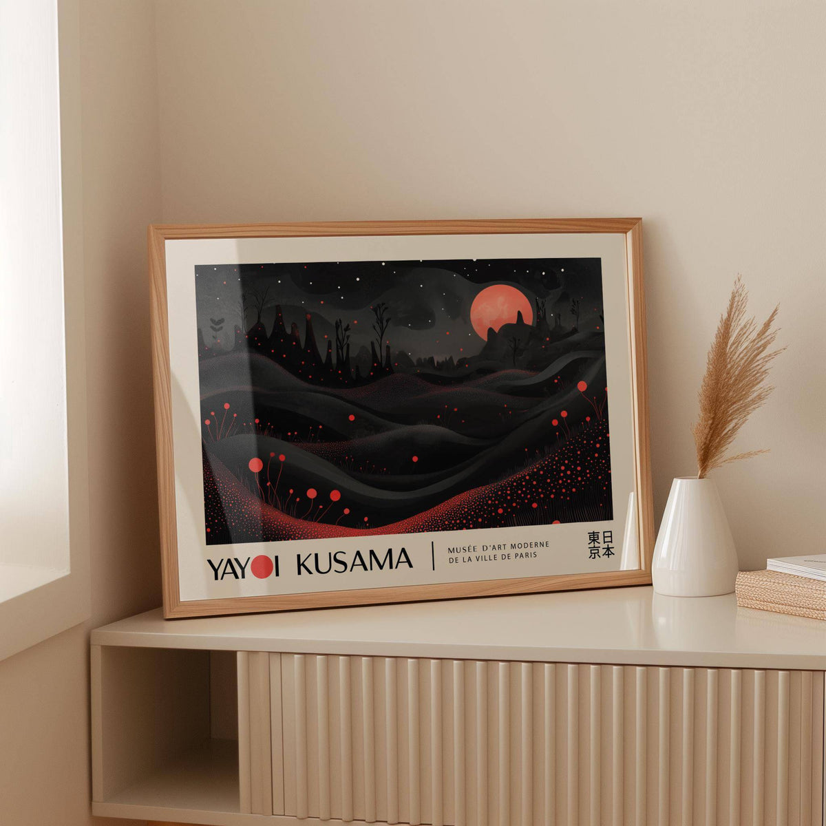 Framed Moon Print with red moon and dots leaning on a beige sideboard next to a white vase with pampas