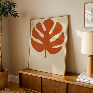 Monstera print in burnt orange leaning on a wooden sideboard in a warm living room