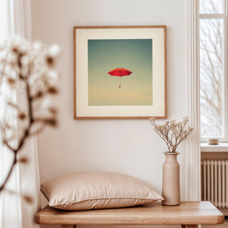 Surreal wall art of a red umbrella floating against a muted sky above a wooden bench with a cushion and vase