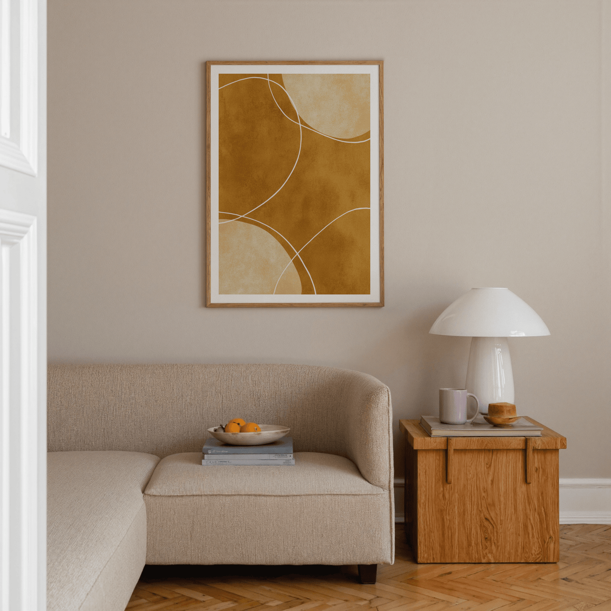 Minimalist geometric art print with golden and beige curved shapes above a beige sofa and wooden side table with a lamp