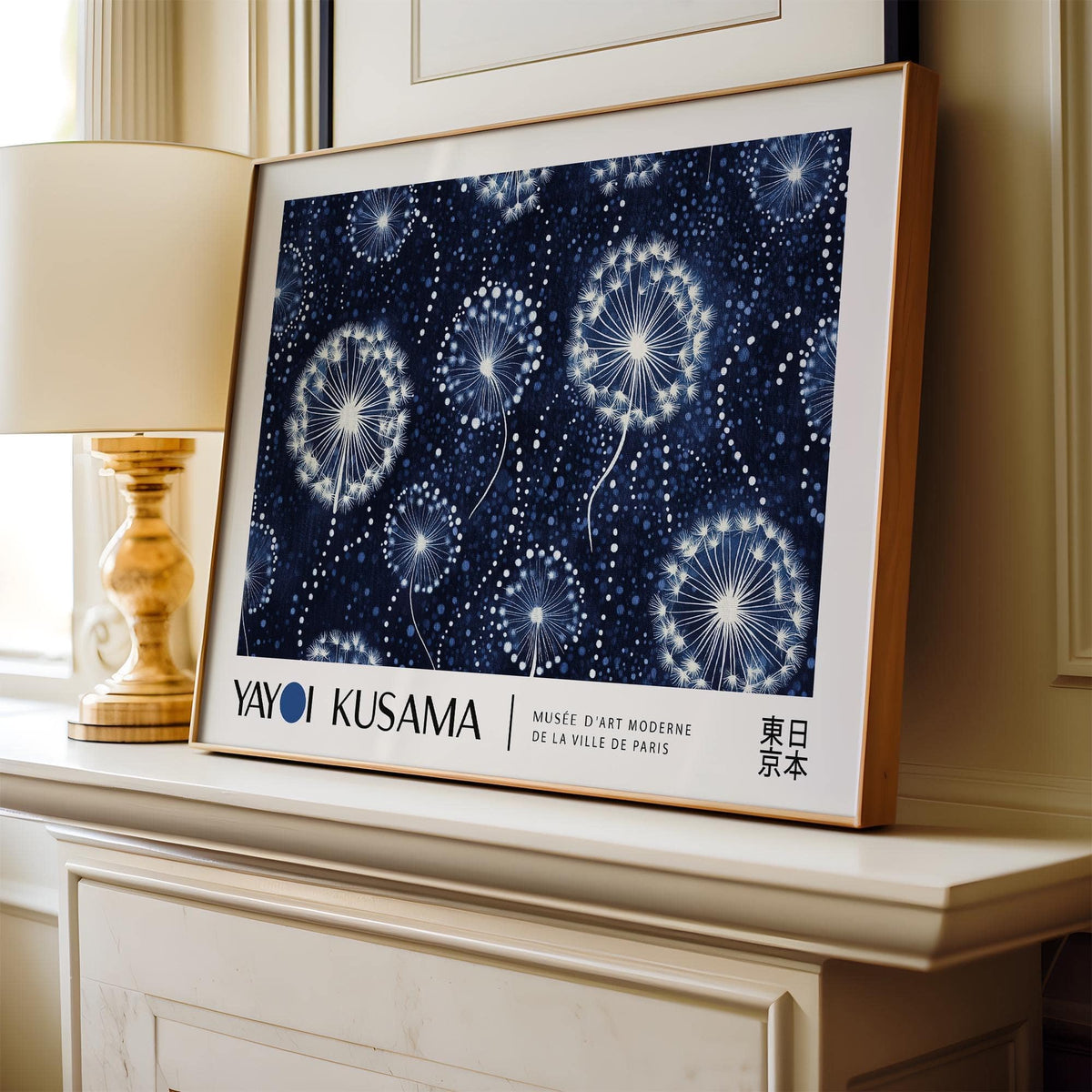 Framed print of dandelion seed heads in white on a dark blue background leaning on a white mantelpiece by a table lamp