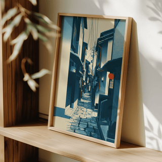 Framed Japanese Street wall art with blue and cream narrow street leaning on a wooden shelf with blurred leaves