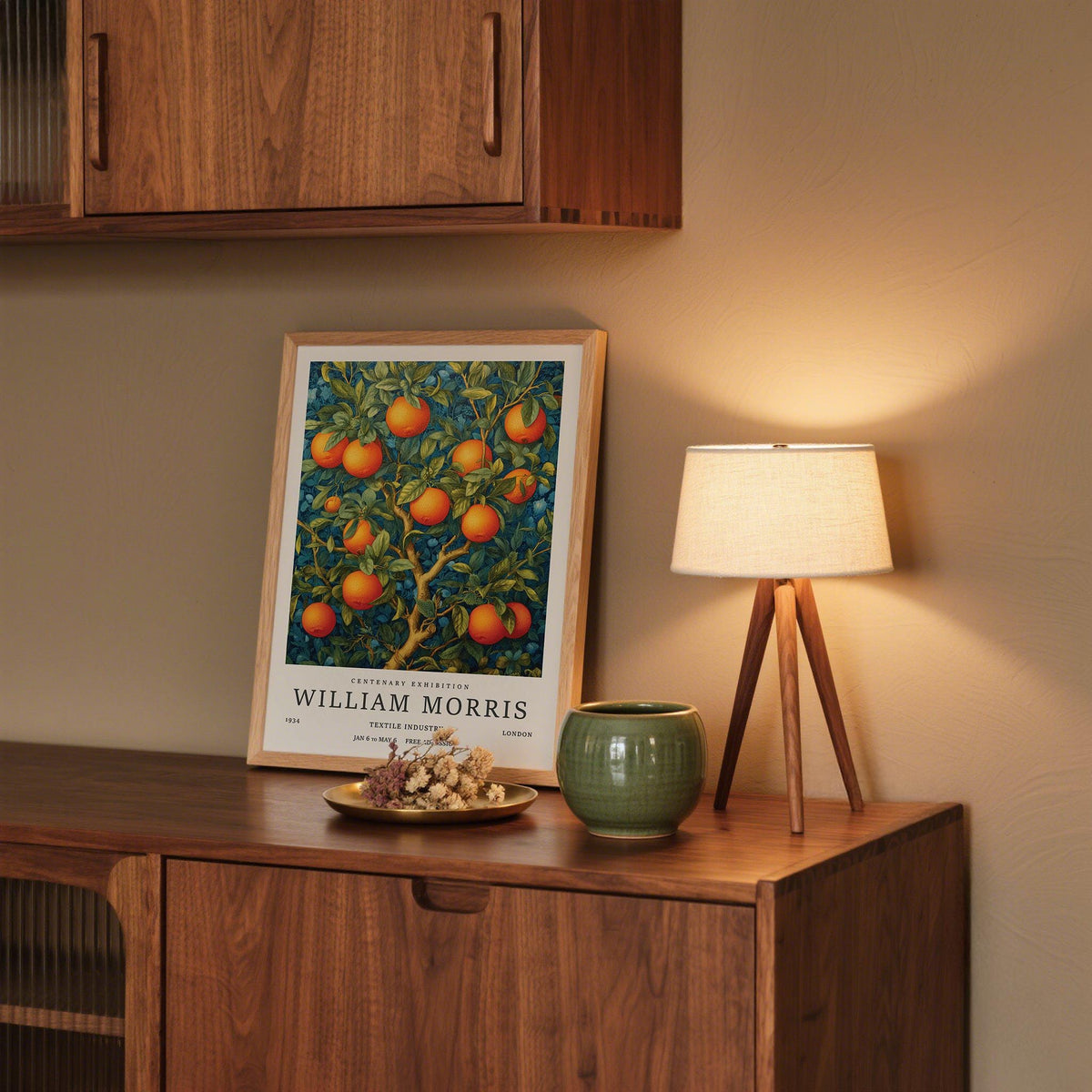 Print of an orange tree with fruit and text leaning on a wooden sideboard beside a small lamp and green pot