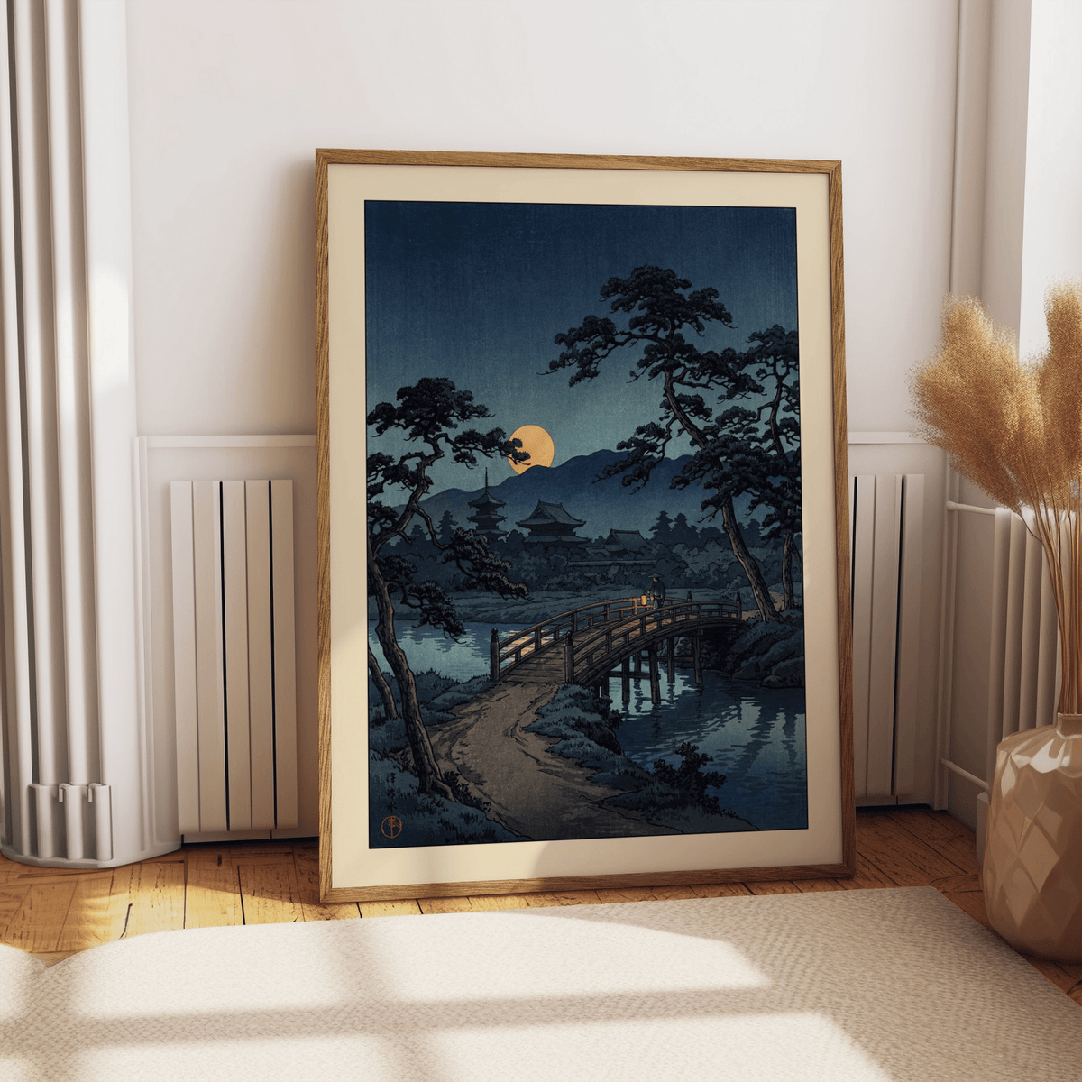 Framed moonlit bridge art print with pine trees and yellow moon leaning on wooden floor by a radiator