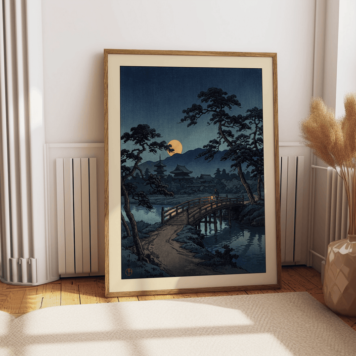Framed moonlit bridge art print with pine trees and yellow moon leaning on wooden floor by a radiator