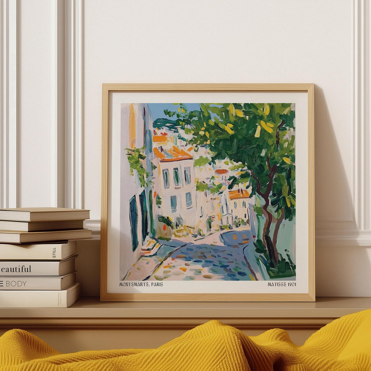 Framed urban street scene print with green tree and colourful houses resting on a shelf with books