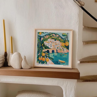 Print leaning on a wooden shelf showing Amalfi Coast Painting with vibrant oranges and blue waters near curved staircase