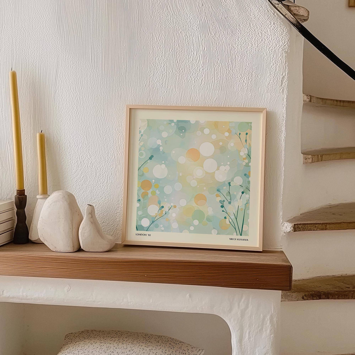 Print leaning on a shelf with abstract circles and dots in soft blue green and beige beside two candles