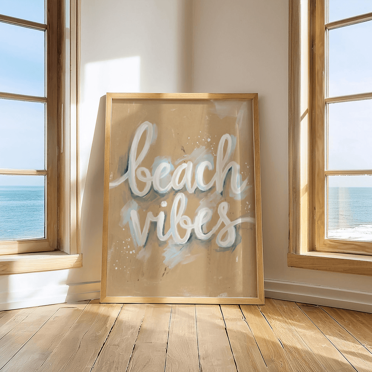 Print on a wooden floor of Beach Vibes wall art with white and blue text between two windows showing an ocean view