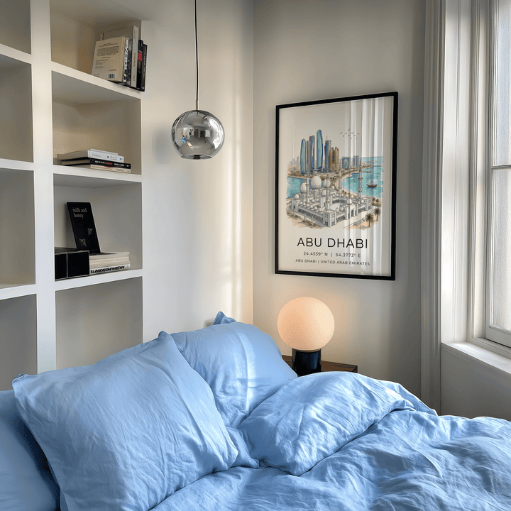 Abu Dhabi Art Print in a black frame showing an illustration of the skyline with blue and beige tones above a lamp in a bedroom