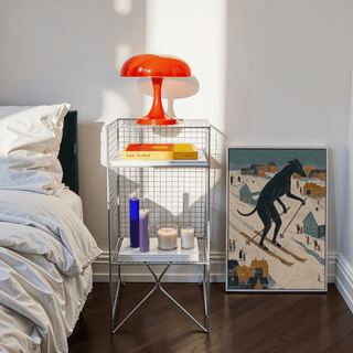 Print leaning on the floor of skiing dog art print in folk-art style with muted blue and orange colours beside a white bed