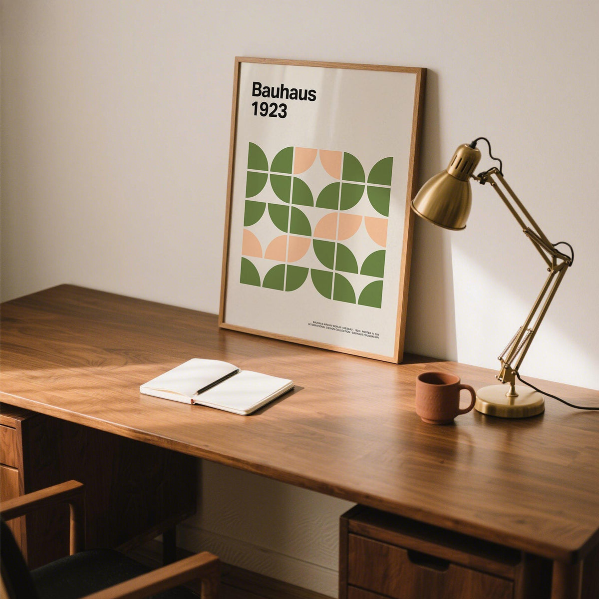 Print leaning on a desk of abstract geometric shapes in green and peach with a gold desk lamp nearby