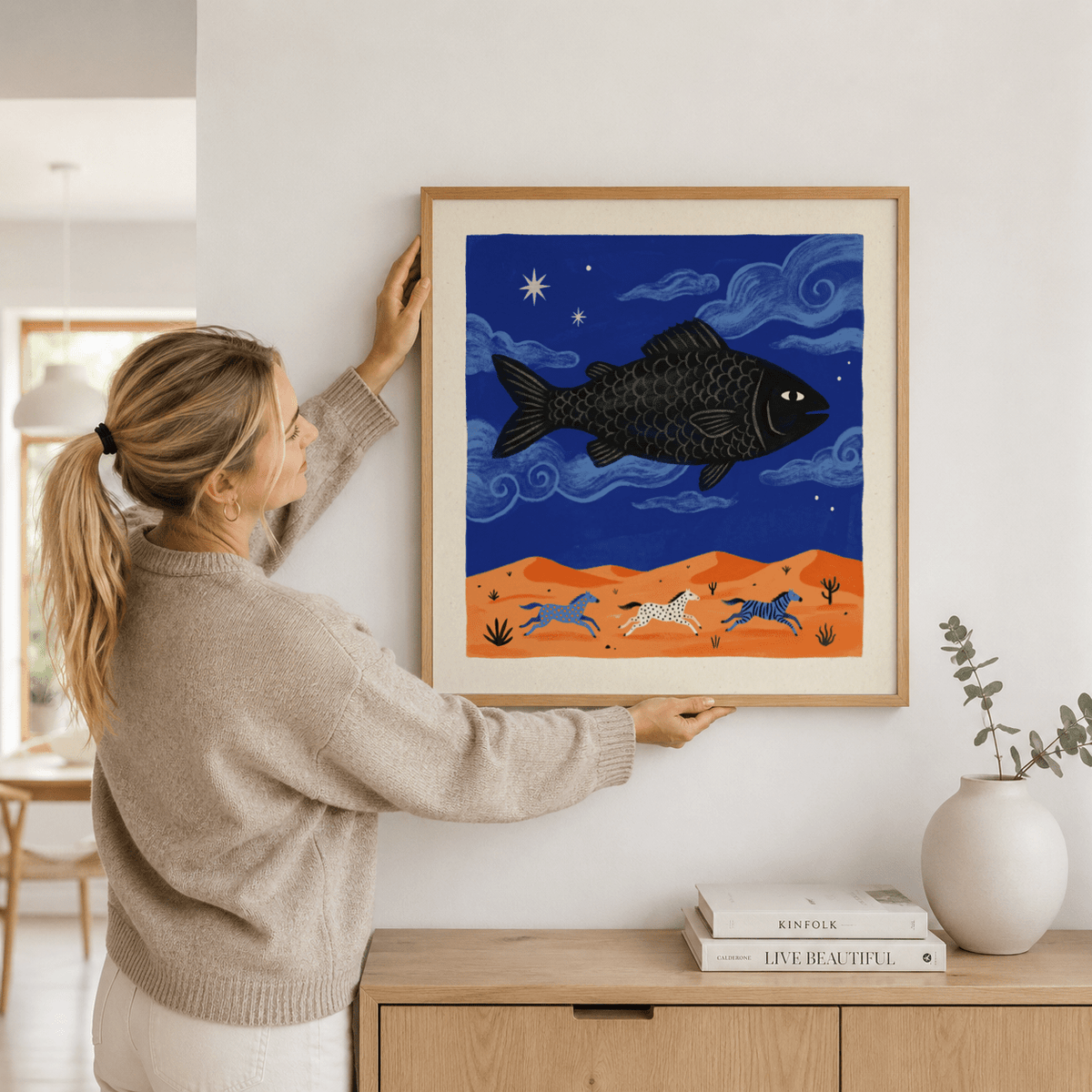 Whimsical Fish Art Print held in hands showing a flying black fish over orange land with three zebras and blue sky