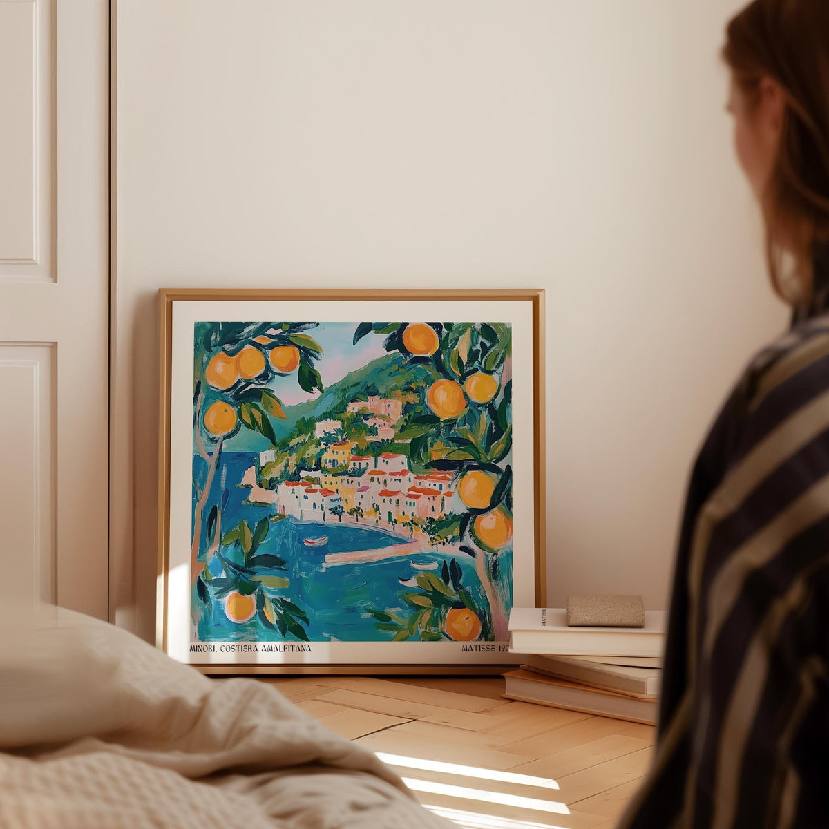 Amalfi wall art leaning on wooden floor beside a pile of books with warm light