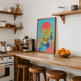 Framed print with yellow flowers and blue leaves on a kitchen counter beside a bowl of oranges