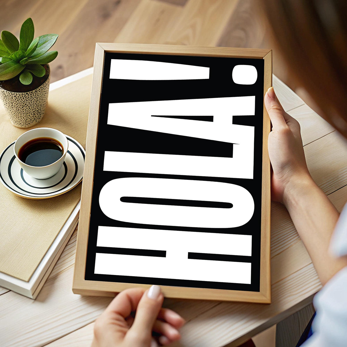 Framed print close-up of bold white HOLA text on black background held in hands above a wooden table