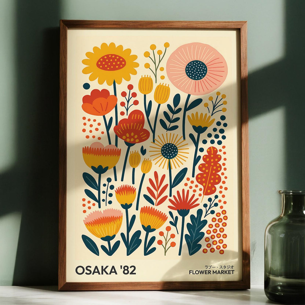 Framed print of wall art flowers with orange yellow and pink stylised blooms and green leaves with a glass vase nearby