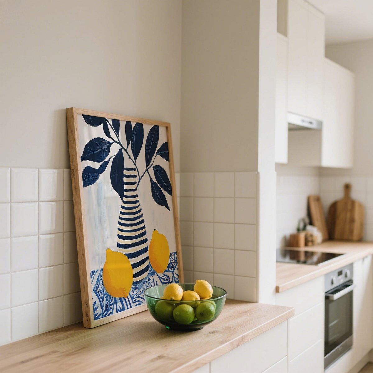 Print leaning on a kitchen counter featuring striped vase with lemons and dark blue leaves beside a bowl of lemons and limes
