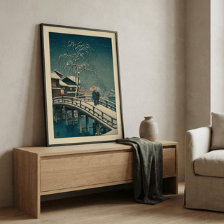 Framed Hasui Kawase Snow Print leaning on a wooden sideboard with a ceramic vase and grey cloth