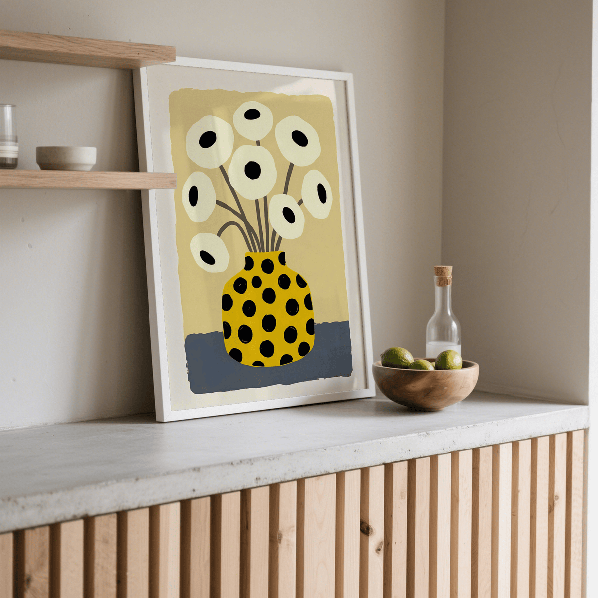 Print leaning on a shelf of yellow vase poster with white flowers and black spots next to a fruit bowl