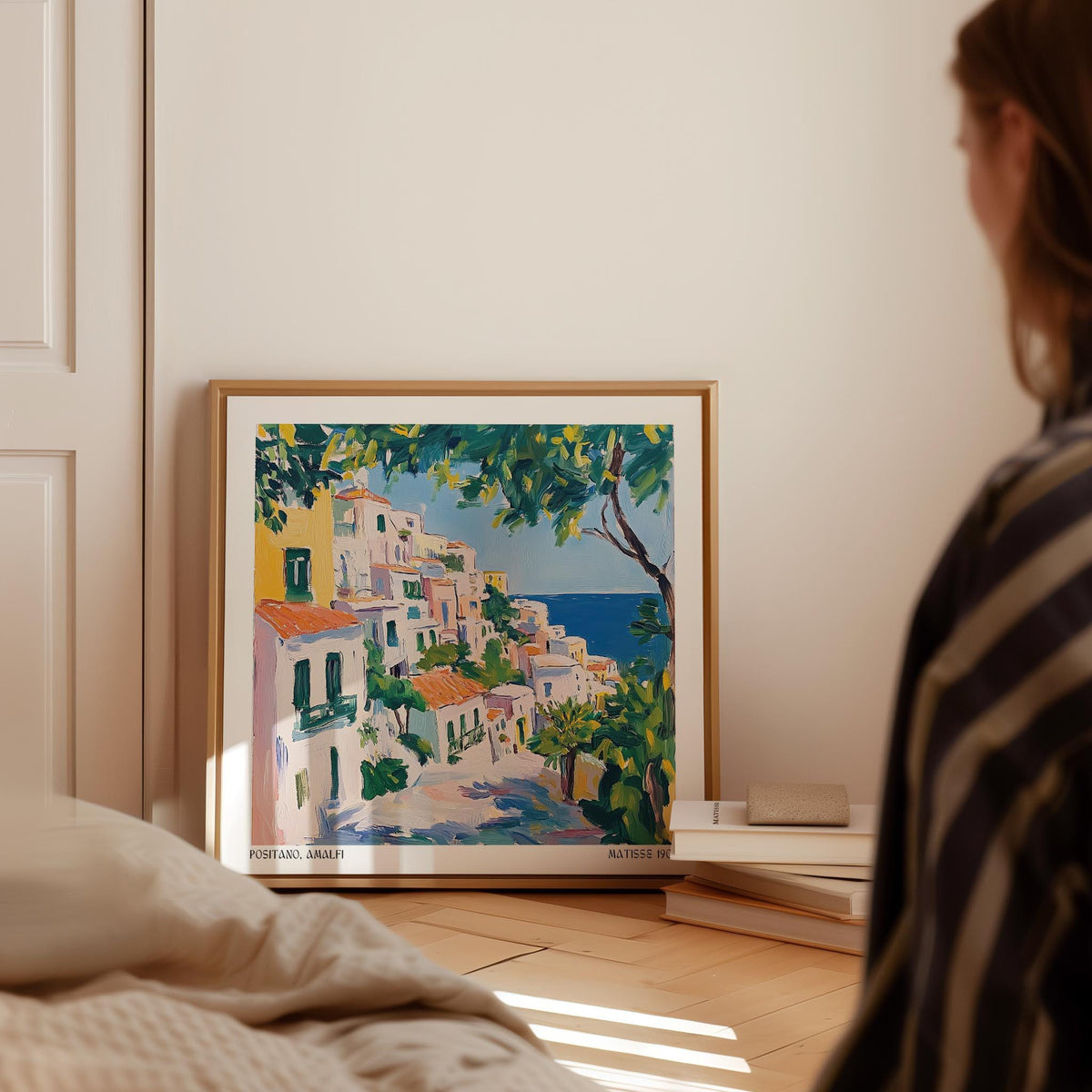Positano Art print leaning on the floor with white bindings near stacked books in a bright room