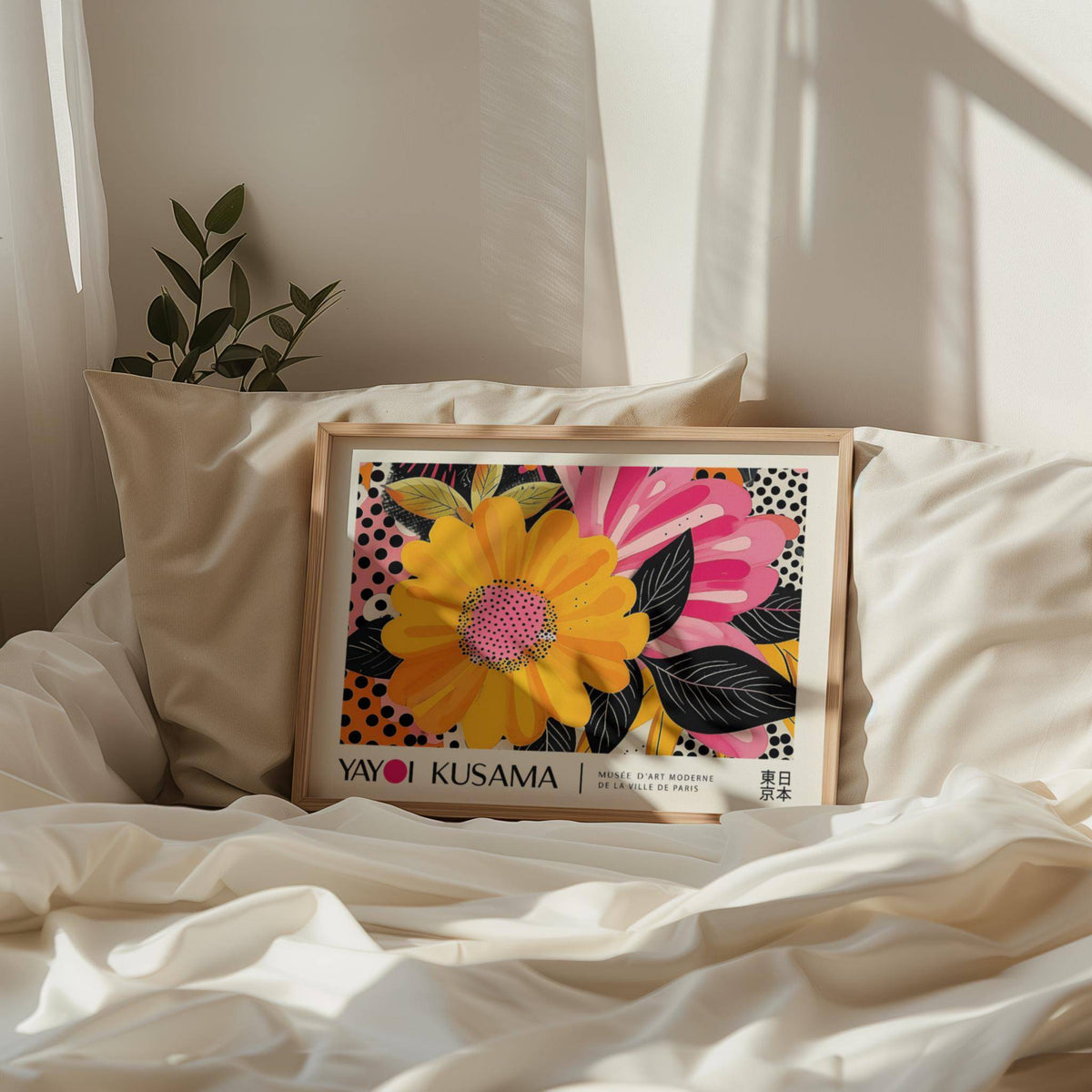 Framed print of bright yellow and pink pop-art flowers resting on crumpled white bedding with a small leafy plant nearby