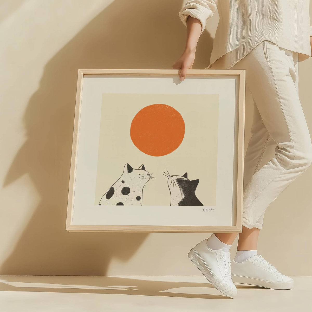 Framed Minimalist Cat Sunshine wall art held by a person showing two black and white cats and an orange circle