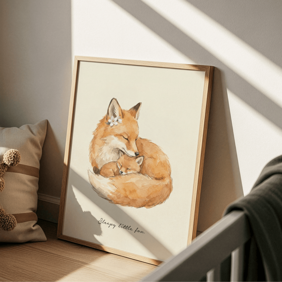 Print leaning on a floor next to a cushion with pom poms featuring watercolour illustration of sleeping mother fox and kit