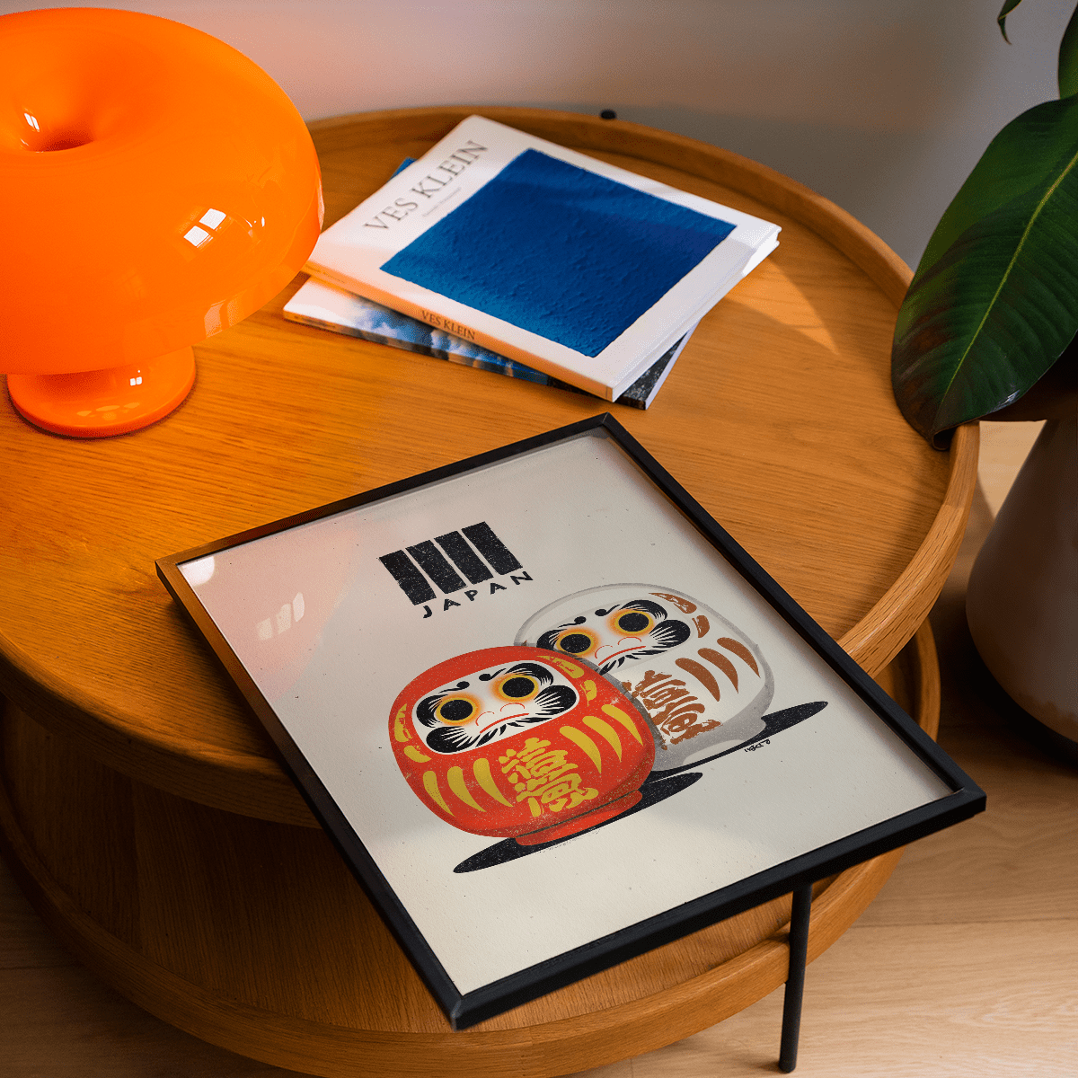 Framed Daruma Print with red and white dolls on a round wooden table next to an orange lamp and green plant