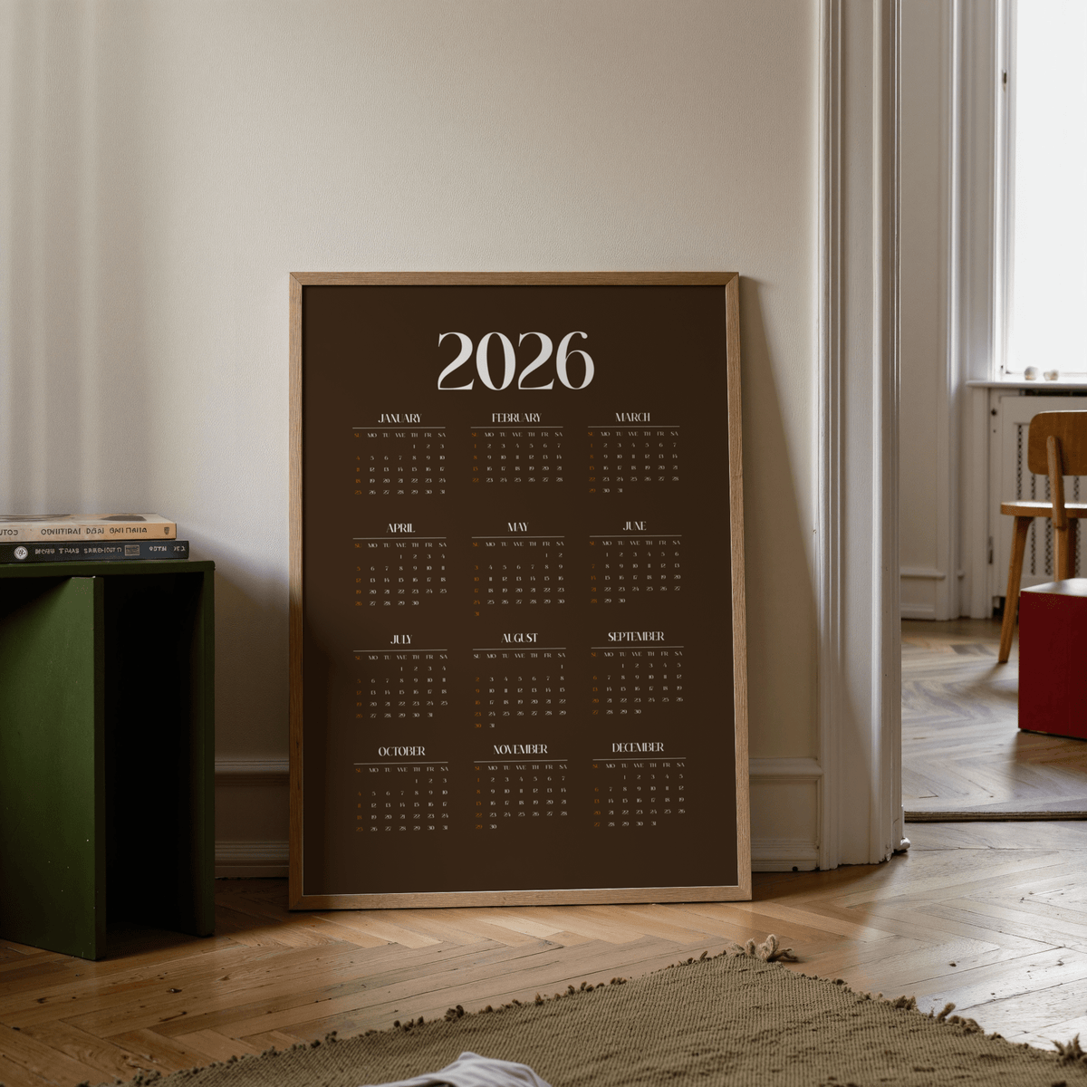 Print leaning on floor showing a minimalist 2026 calendar in brown with a wooden frame beside a green shelf
