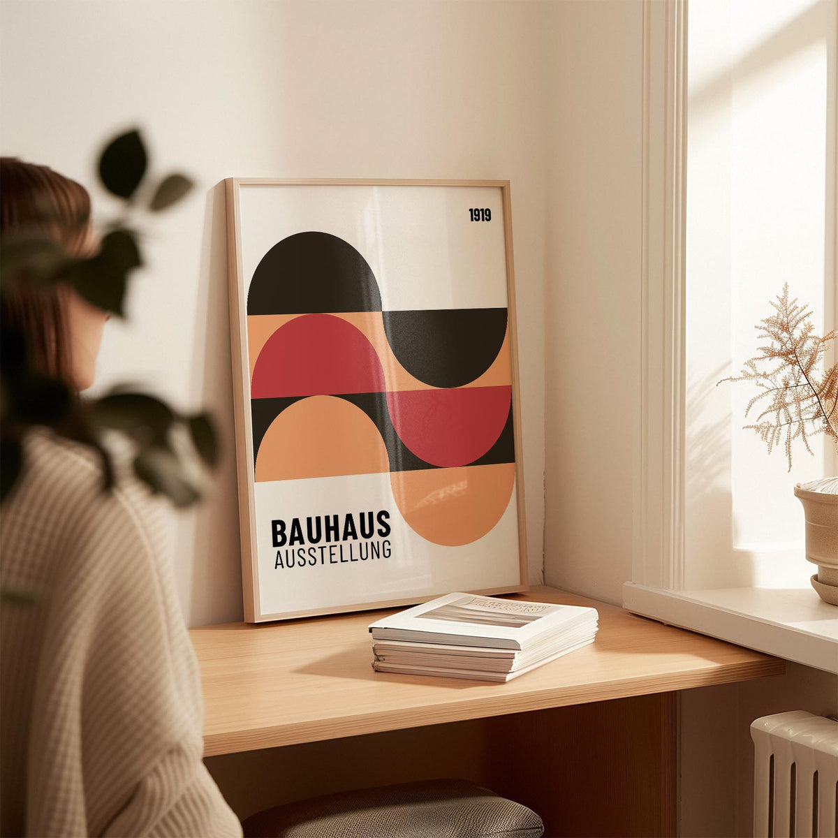 Print leaning on a desk of abstract geometric shapes in red black and beige next to books and a plant