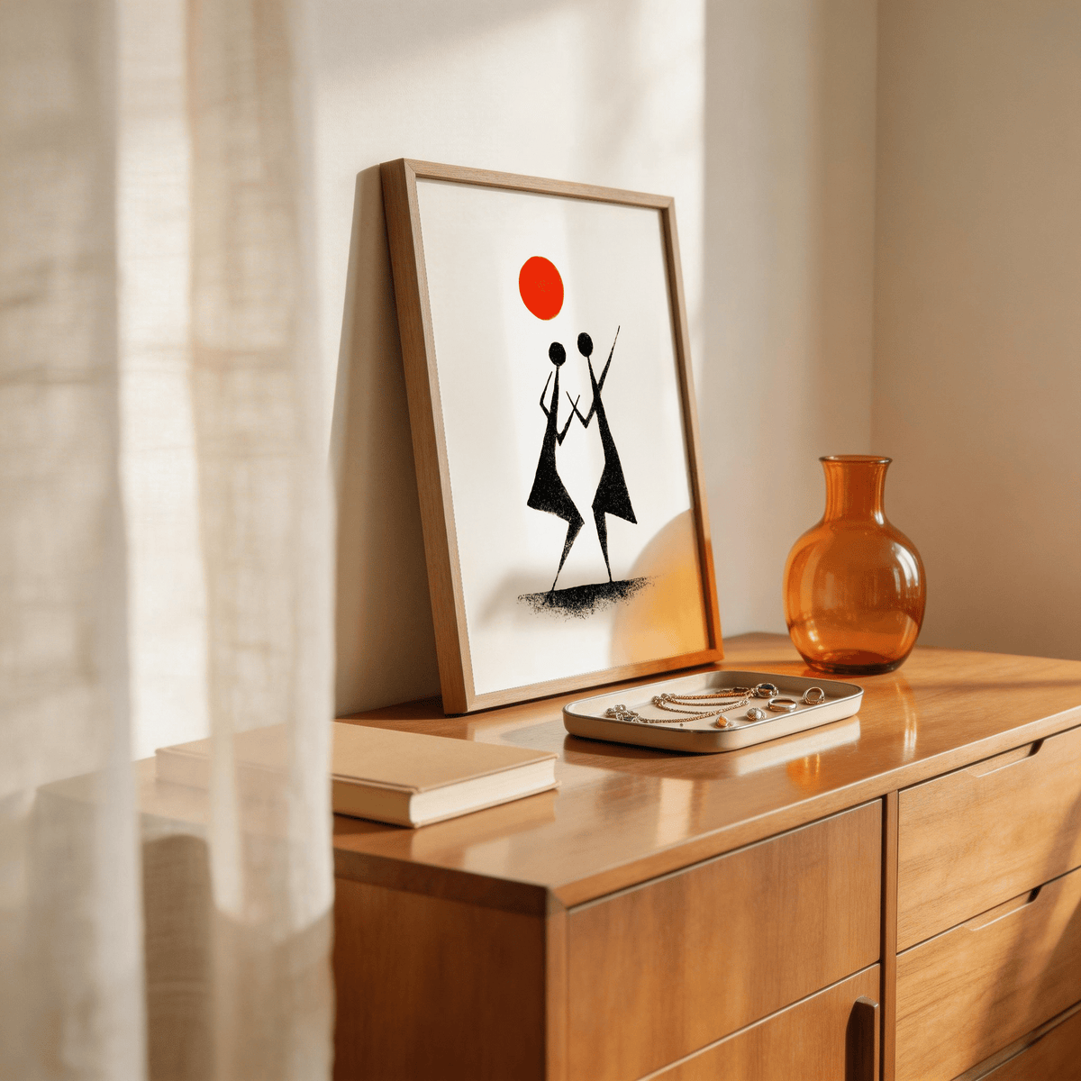 Framed print of Warli Folk Art with black tribal dancers and an orange sun leaning on a wooden dresser with a glass vase