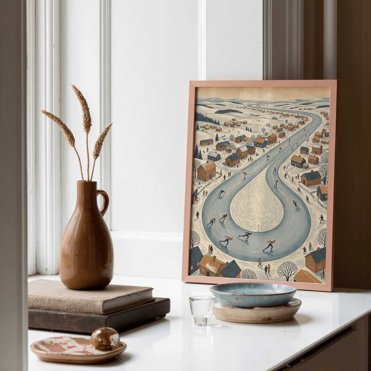 Winter Village Scene print leaning on a white surface with brown pottery and books nearby in a neutral room