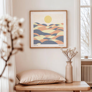 Pastel wall art of abstract hills with sun in soft blue, beige and peach tones above a wooden table with vase and cushion