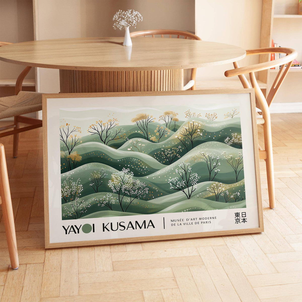 Landscape art prints leaning on a wooden floor with minimalist hills and blossoming trees in soft greens and yellows beside a wooden table