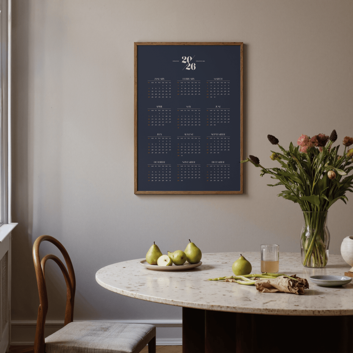 Minimalist Calendar print with white and orange text on navy above round table with pears in a dining room
