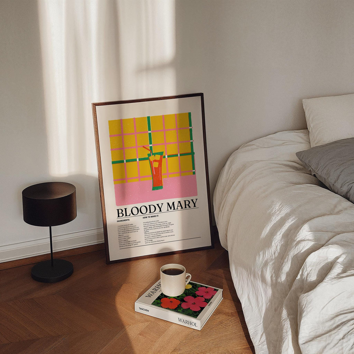 Print leaning on a floor beside a bed showing a Bloody Mary cocktail with yellow and pink grid background