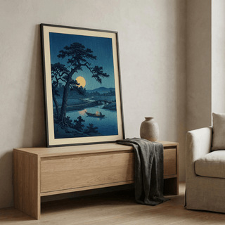 Framed moonlight print of a Japanese woodblock moonlit river scene leaning on a wooden cabinet with a vase