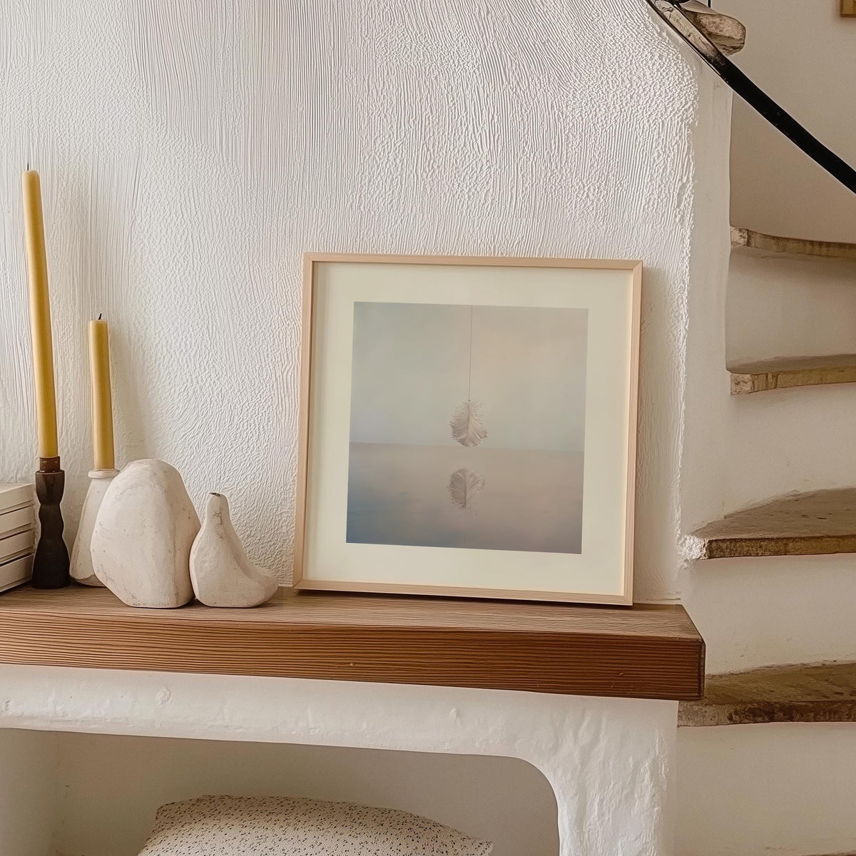 Print leaning on a shelf showing a minimalist white feather with soft blue and beige tones near a staircase and clay decor