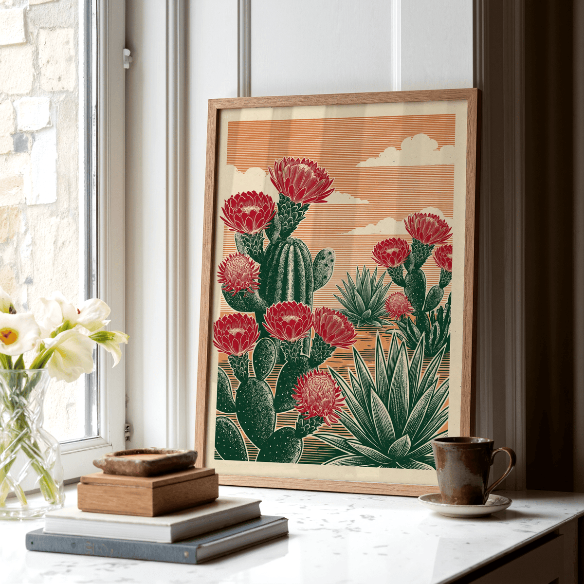 Print leaning on a windowsill of cactus desert art with red flowers and green plants alongside a coffee cup