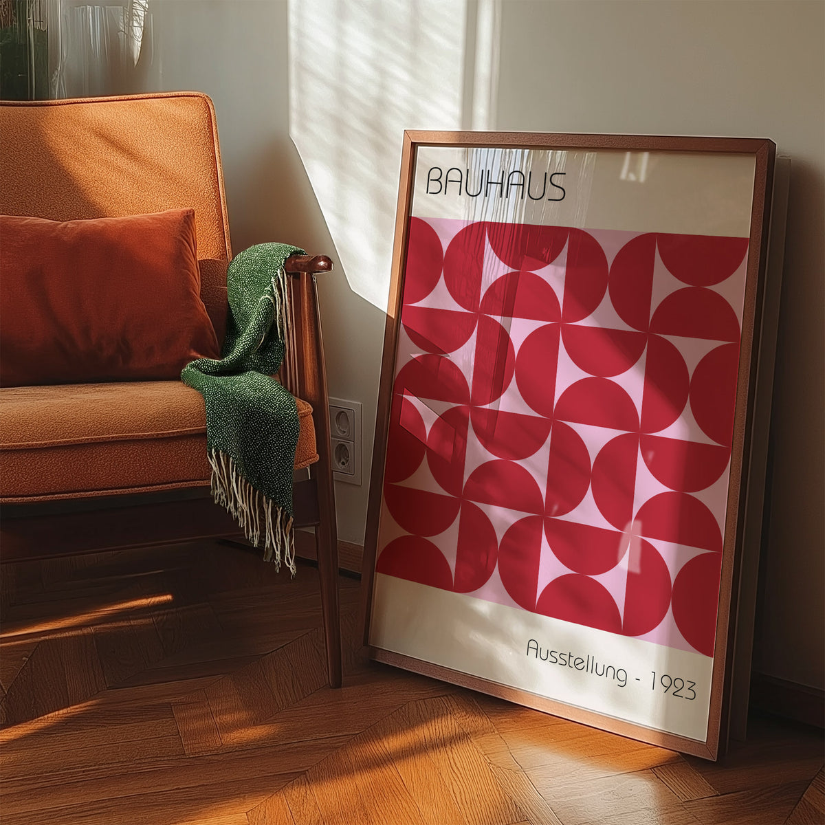 Bauhaus artworks print leaning beside a chair with an orange cushion and green throw under warm sunlight