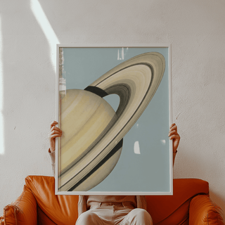 Framed Saturn wall art of minimalist illustration in pale yellow and blue held by a person above a tan sofa