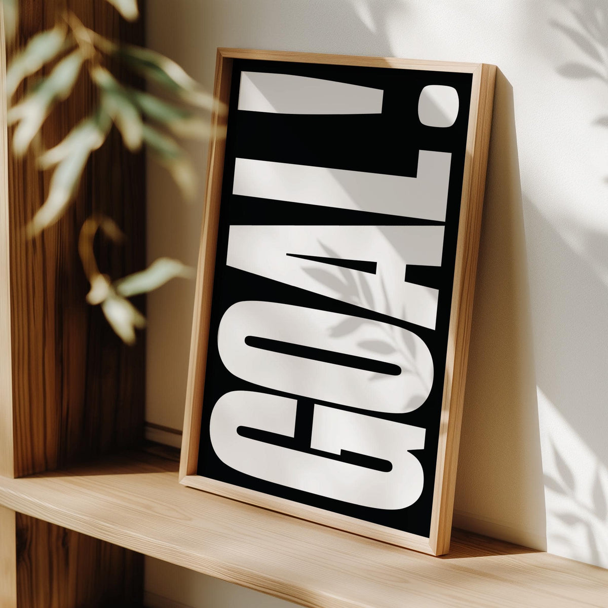 Framed close-up of Goal Poster with large white typography on black leaning on wooden shelf with plant blurred